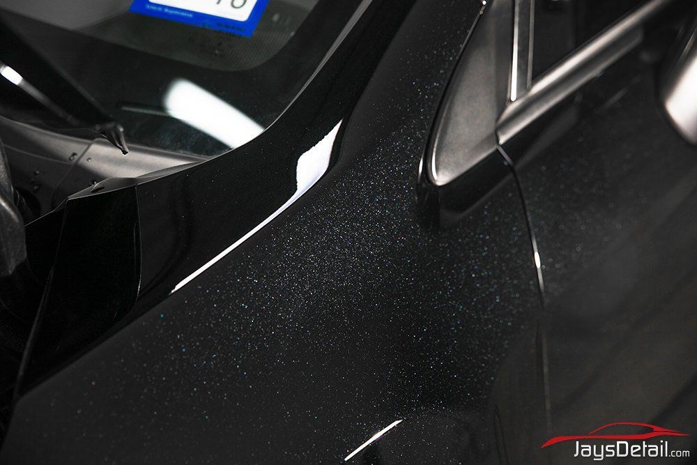 Close-up of a black car's fender, showing speckled paint and a portion of the windshield with a sticker.