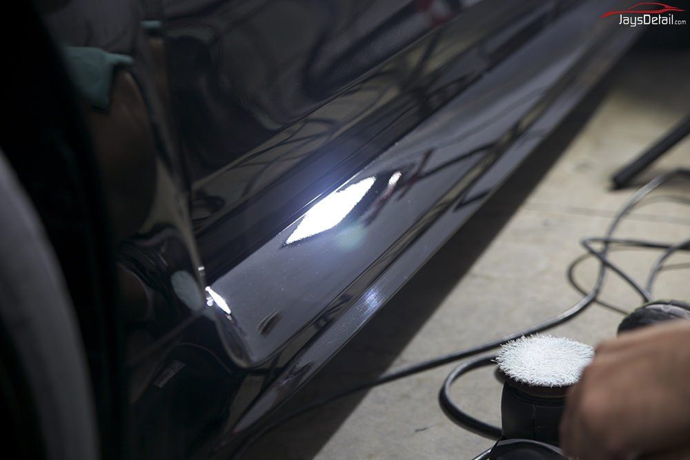 Person polishing a shiny black car panel with a buffing machine; indoor setting.