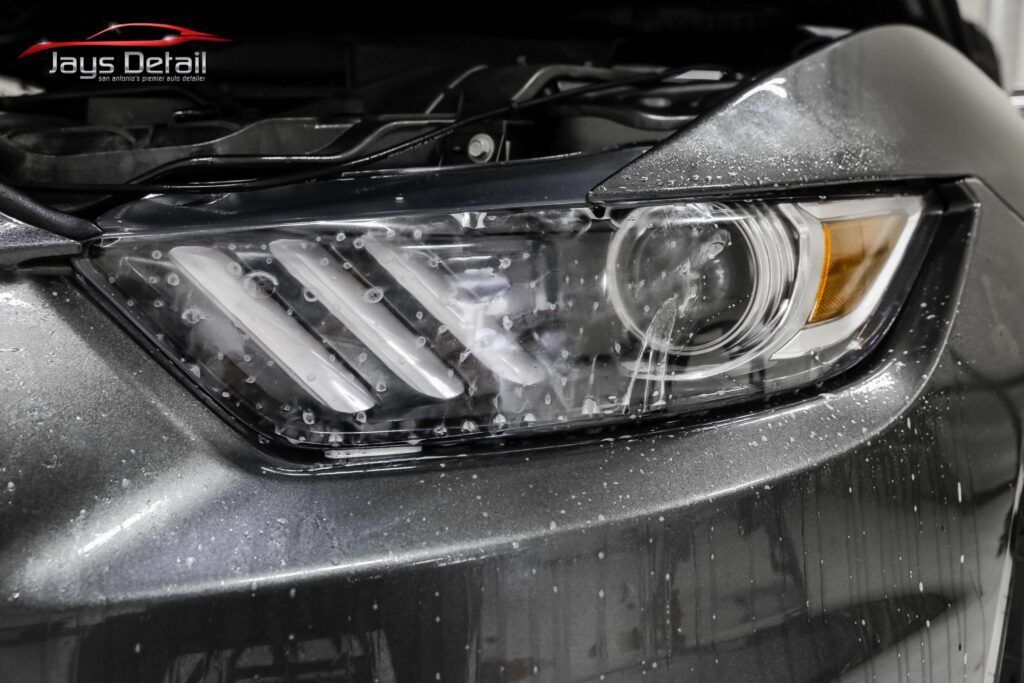 Close-up of a wet, dark gray car headlight. Water droplets glisten on the surface.