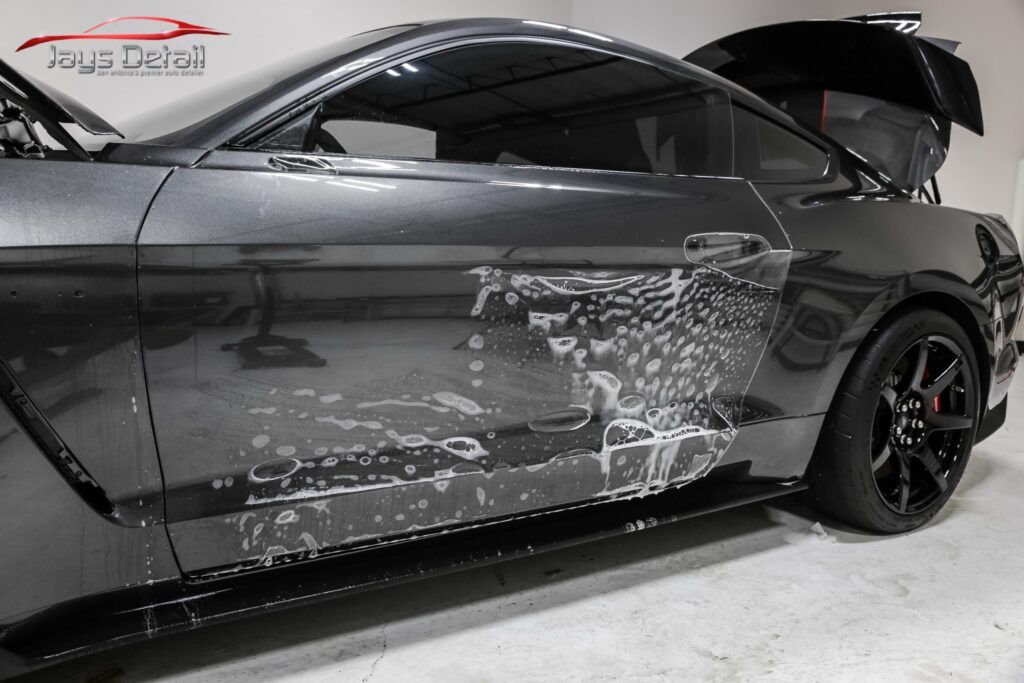 A gray Mustang with soapy film being detailed in a garage. Door is open, and trunk is up.