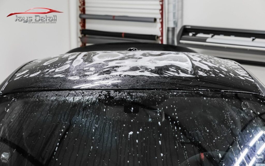 A black car roof is covered in soapy foam, preparing for detailing in a garage.