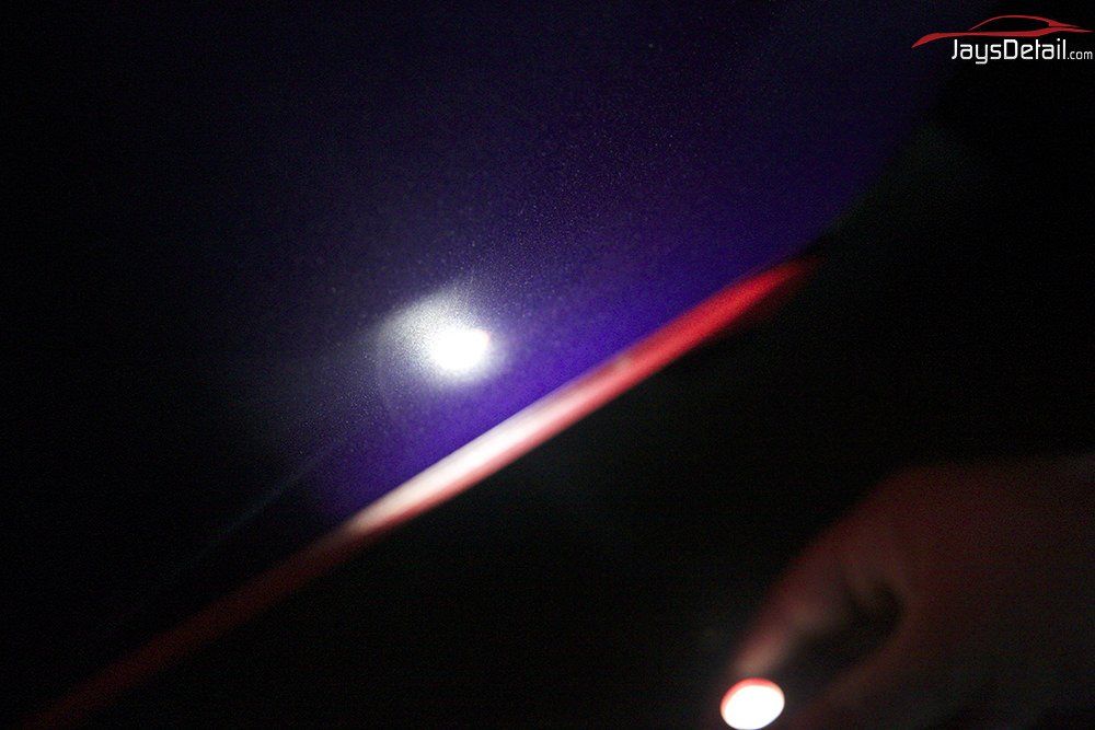 Hand holding a flashlight illuminates car's dark purple paint, revealing a blemish.