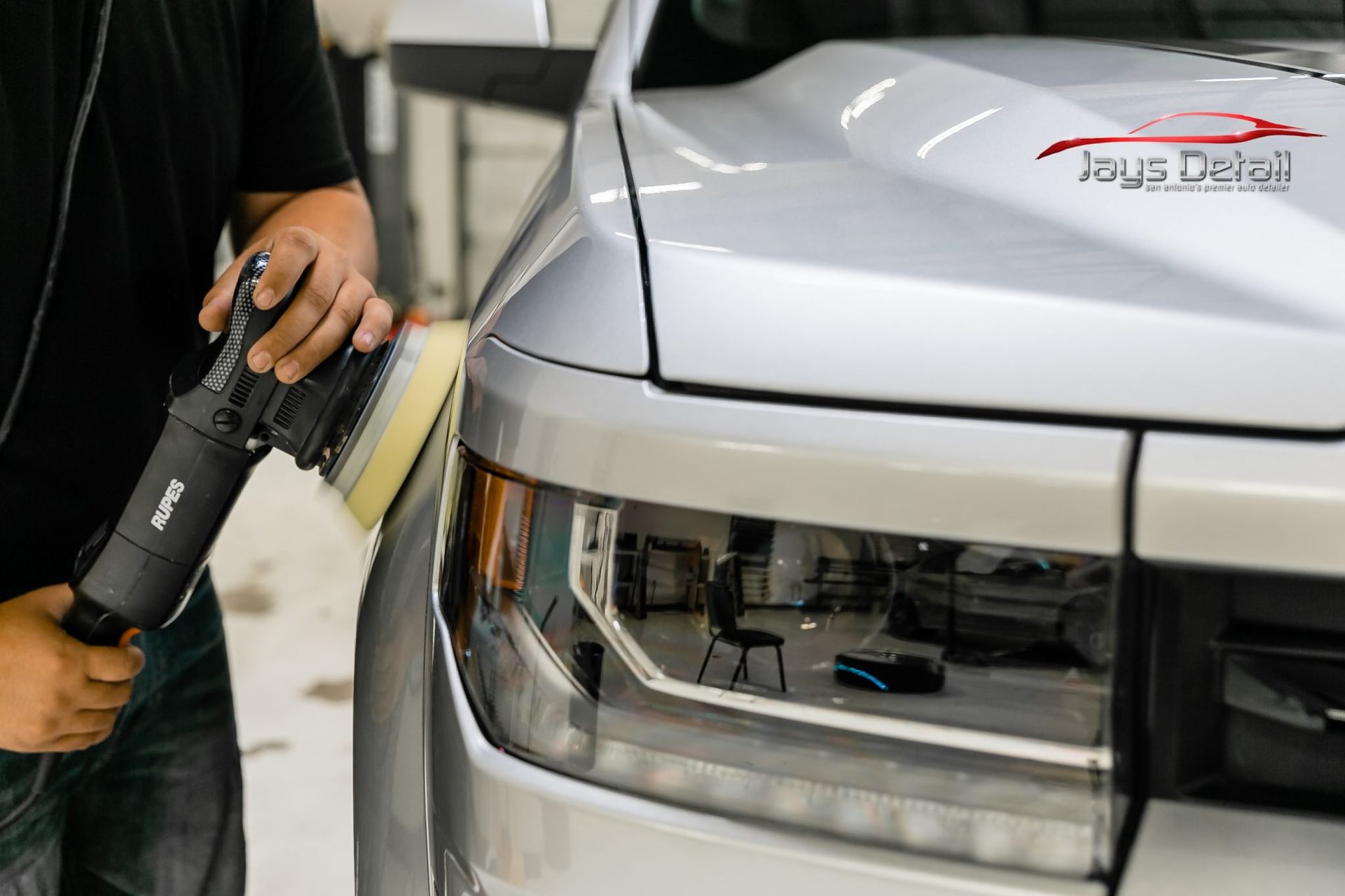 Person polishing the front of a silver truck with an orbital buffer in a garage.
