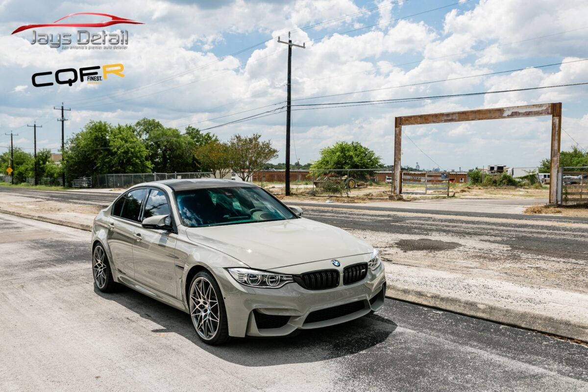 A light gray BMW M3 car parked on a road with a cloudy sky in the background.