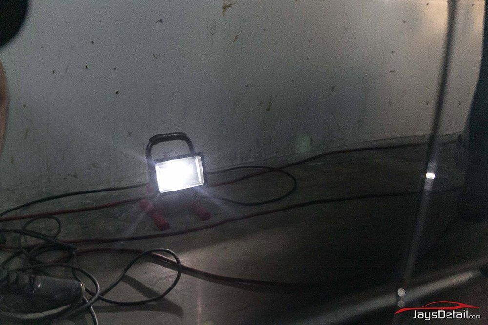 Bright work light illuminates a dark garage floor. A car's side panel is visible.