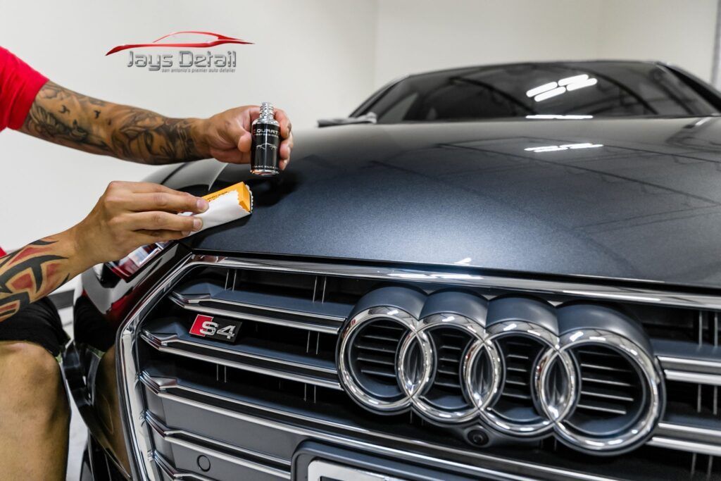 Person applying ceramic coating on a dark Audi S4 hood with chrome grill in a shop.