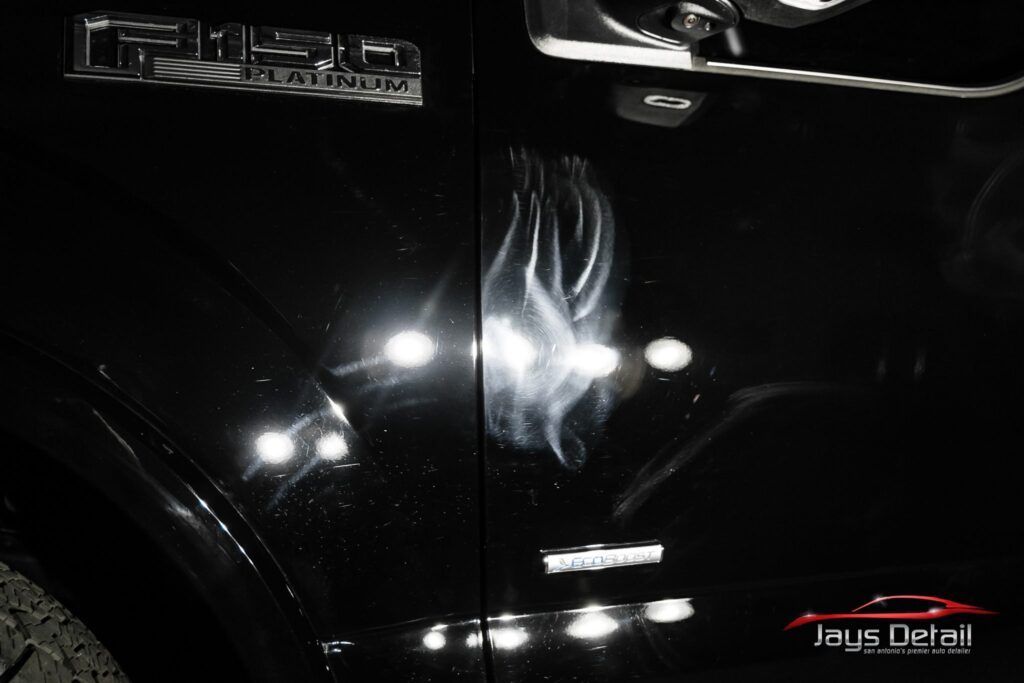 Black truck door with a visible dent and swirl marks reflecting light.