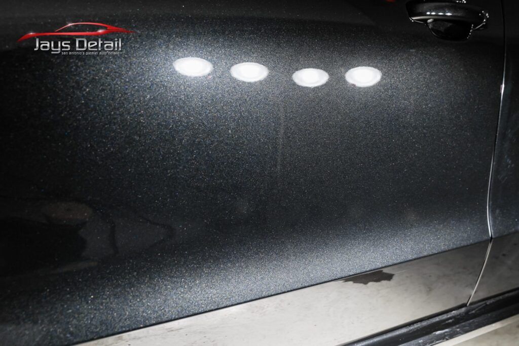 Car door with scratches, reflecting overhead lights. Black paint.