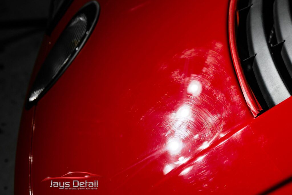 Red car with paint scratches and swirls in a detailing setting.