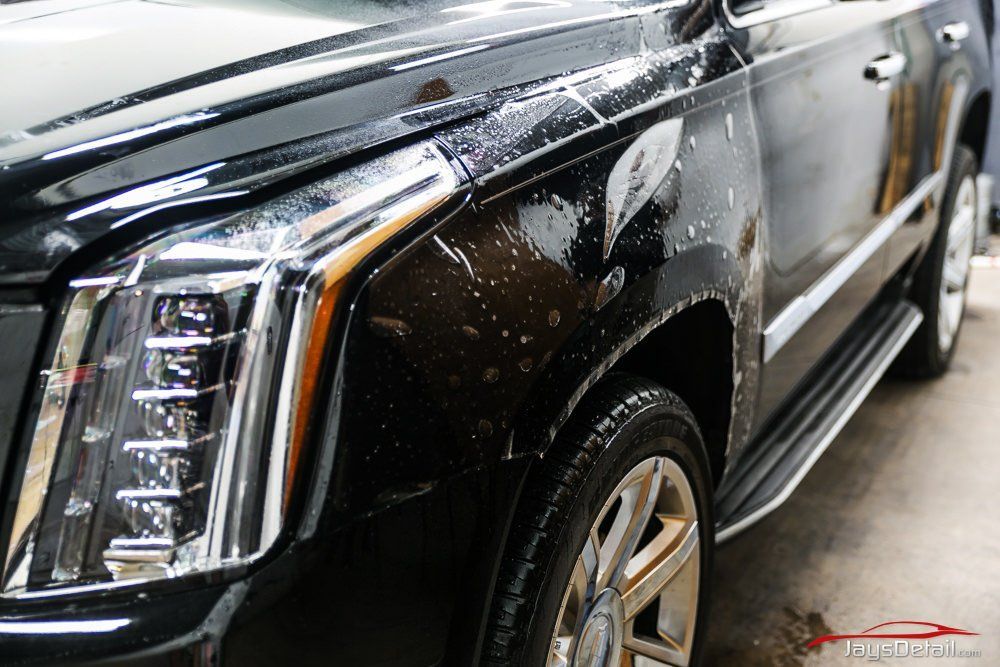 Black Cadillac SUV with clear protective film on the front fender and hood, in a detail shop.