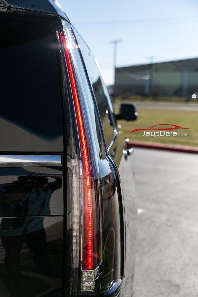 Black Cadillac SUV's vertical taillight illuminated, reflecting a person and sky.
