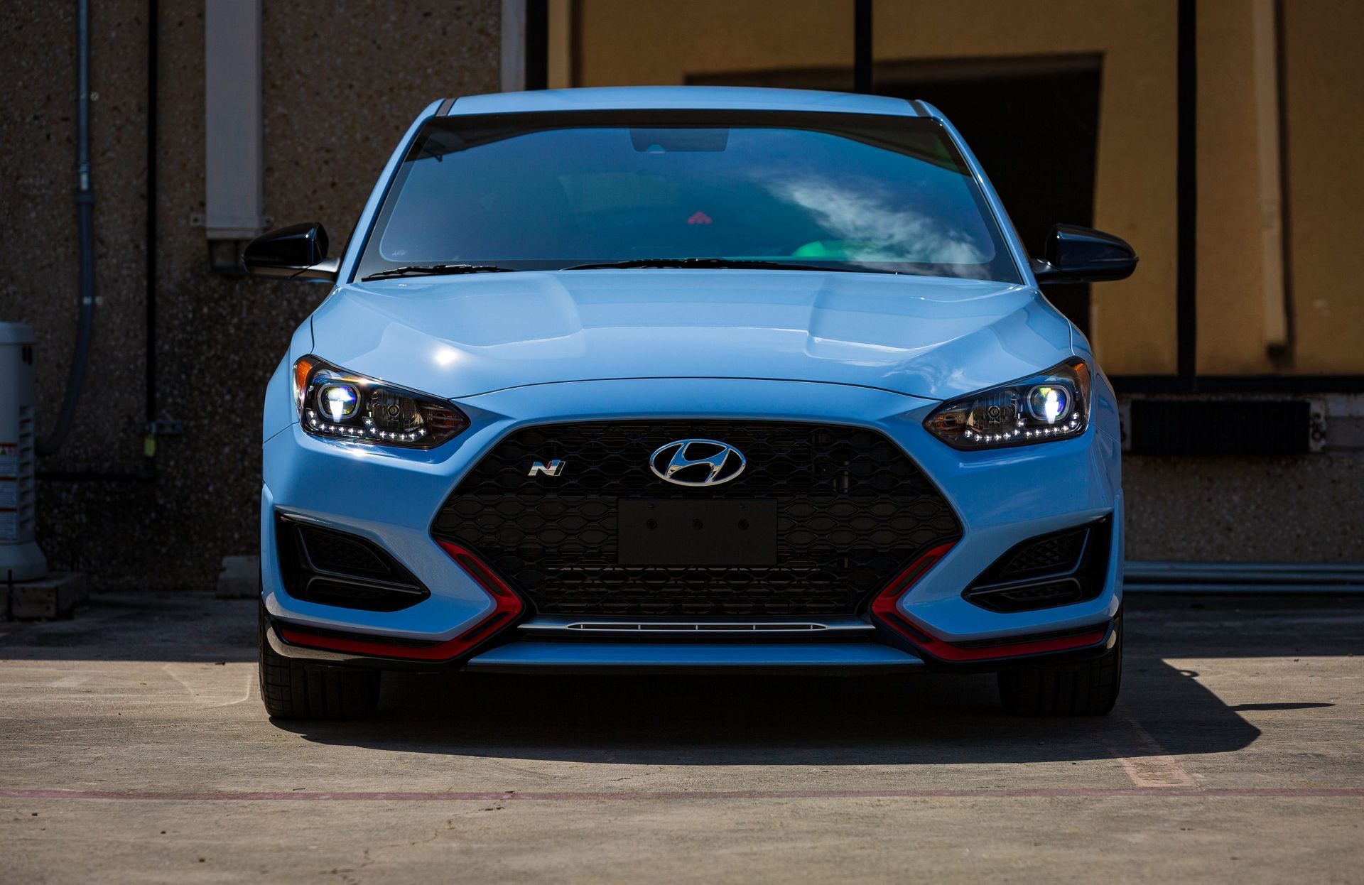 Light blue Hyundai Veloster N sports car, parked in front of a building. Black grill, red accents.