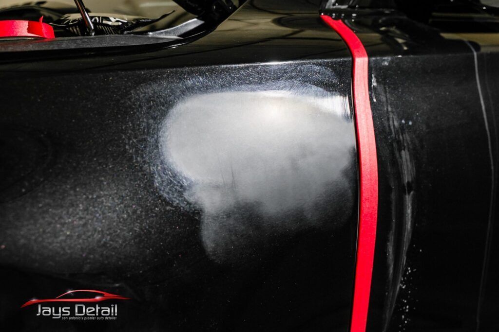 Black car panel with a sanded repair spot, masked with red tape, showing prep work.