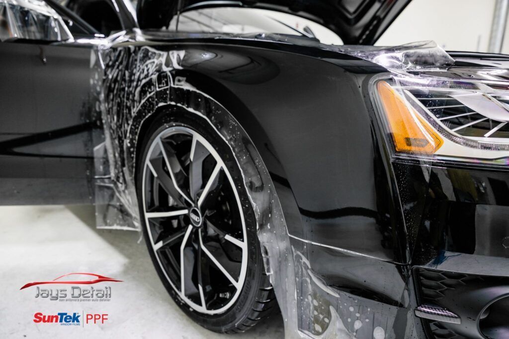 Black car being wrapped in protective film, near a wheel, in a detail shop.