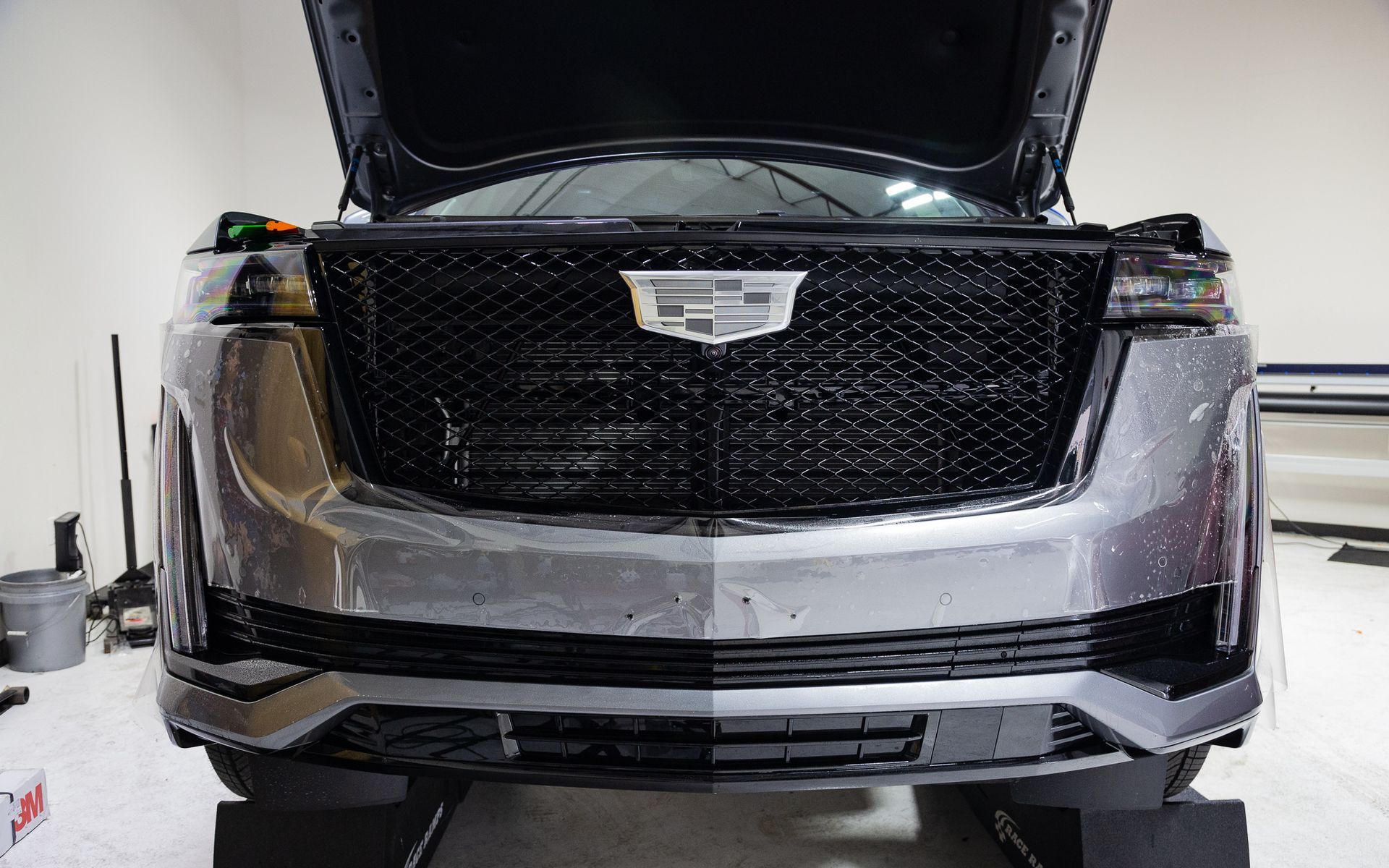 A gray Cadillac SUV front bumper with the hood open. The black grille and the vehicle are on blocks.