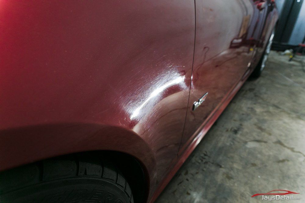 Red car's side panel with reflective surface, showing the wheel, badge, and part of a tire in a workshop.