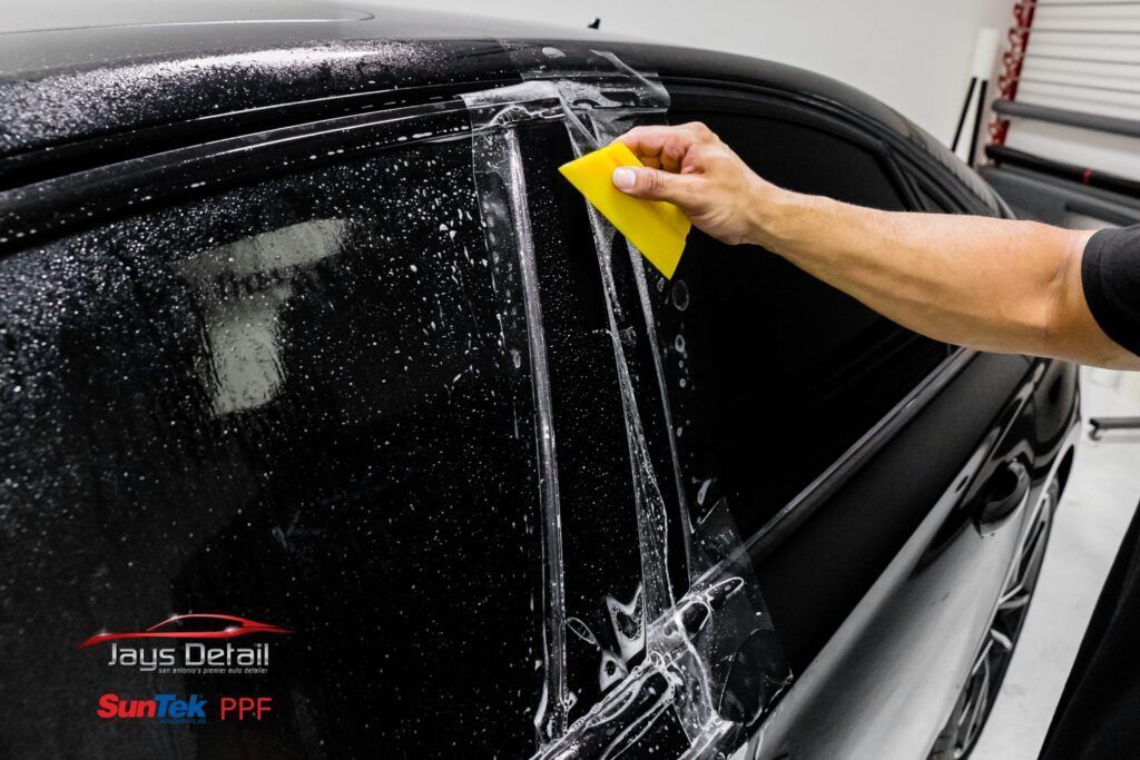 A person applying tint film to a car window using a yellow squeegee in a shop.