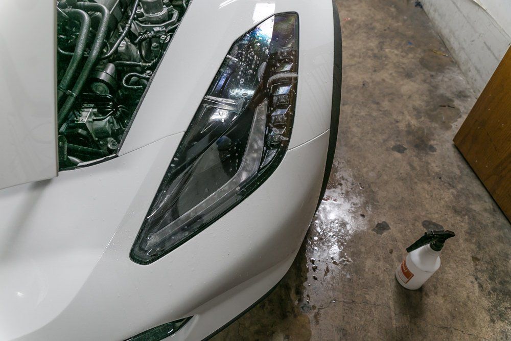 White Corvette's headlight being cleaned with a spray bottle in a garage; wet pavement and open hood.