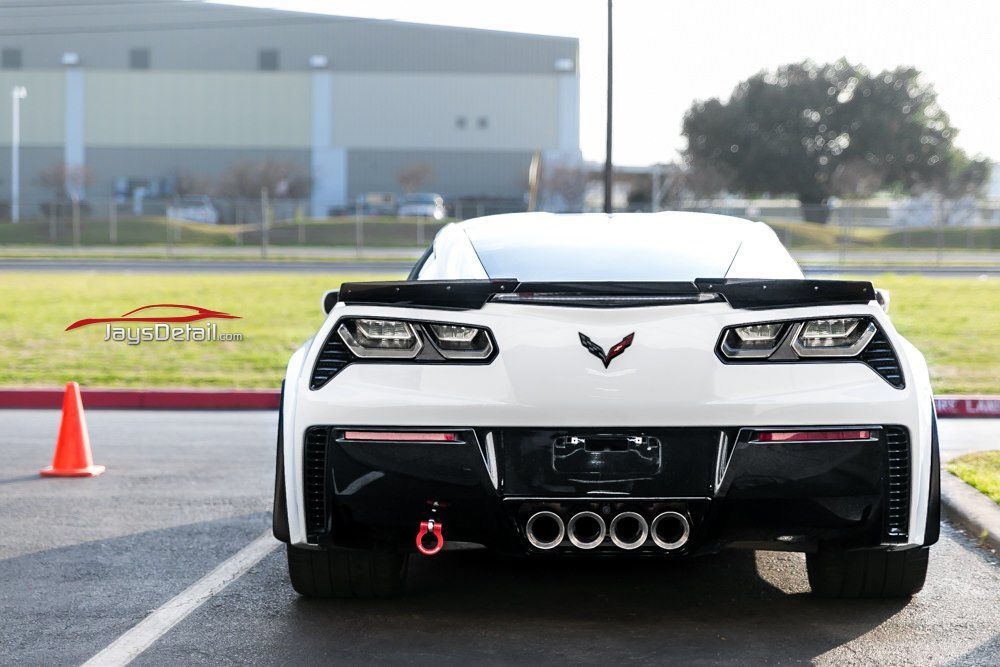 White sports car (Corvette) rear view; black trim, quad exhaust, tow hook, parked outdoors.