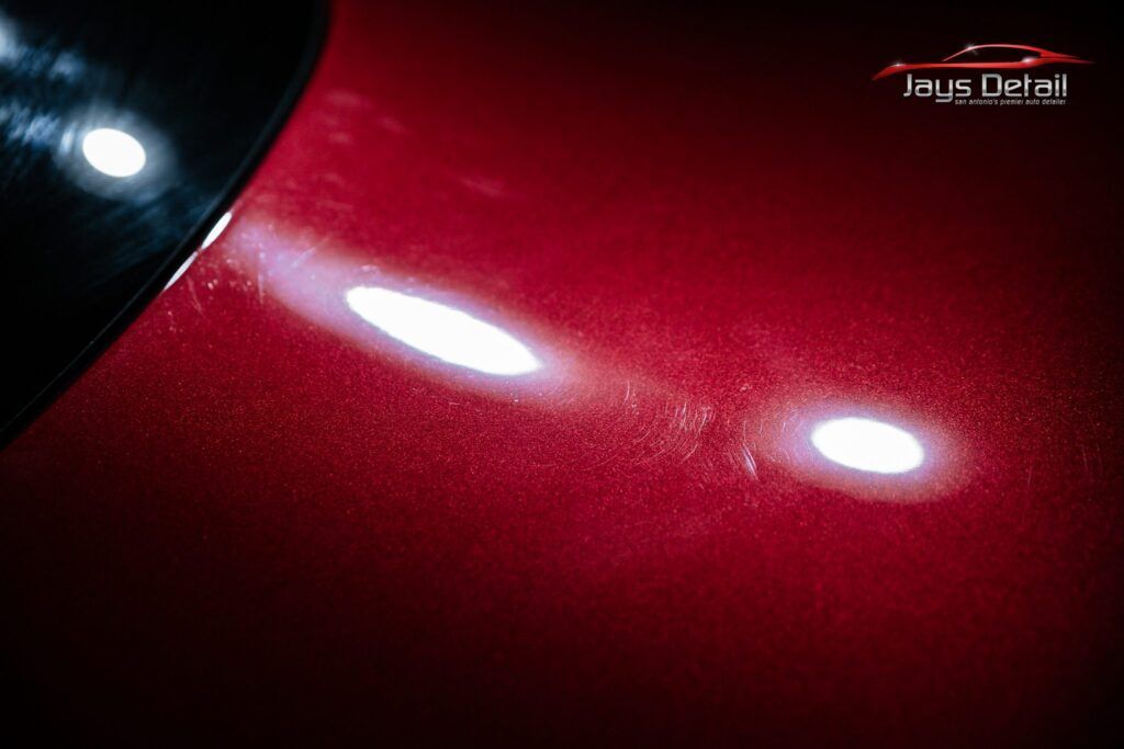 Red car paint with swirl marks illuminated by a bright light, indicating scratches and imperfections.