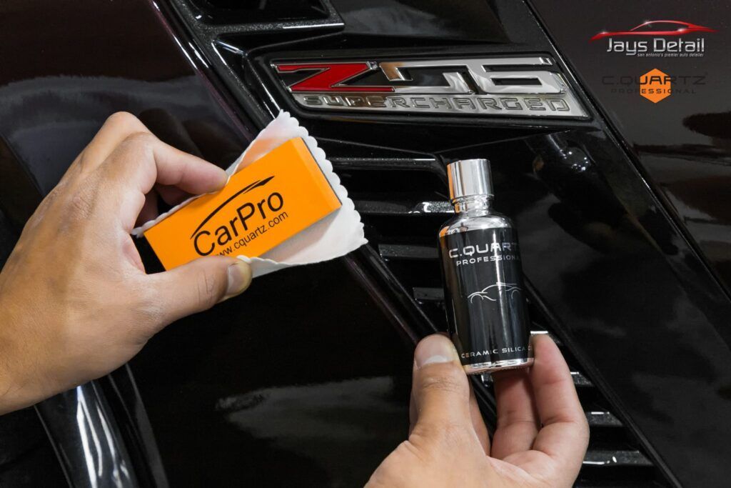 A person applying ceramic coating to a car's emblem with an applicator and bottle, in a black setting.