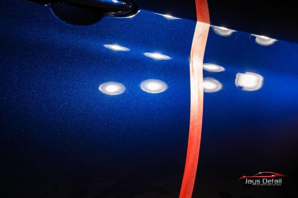 Blue car door with a polished side and a scratched side, separated by red tape, under bright lights.