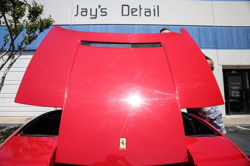 Red Ferrari hood open at Jay's Detail. Bright sunlight reflects off the shiny red paint.