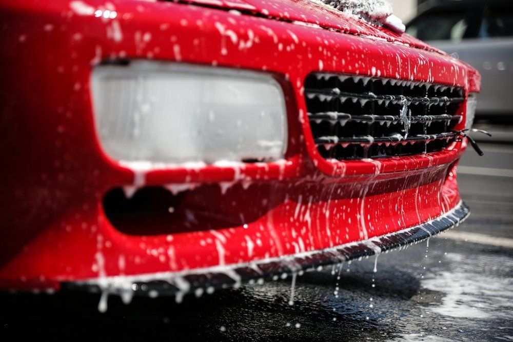Red car being washed; foamy soap on front bumper and headlight.