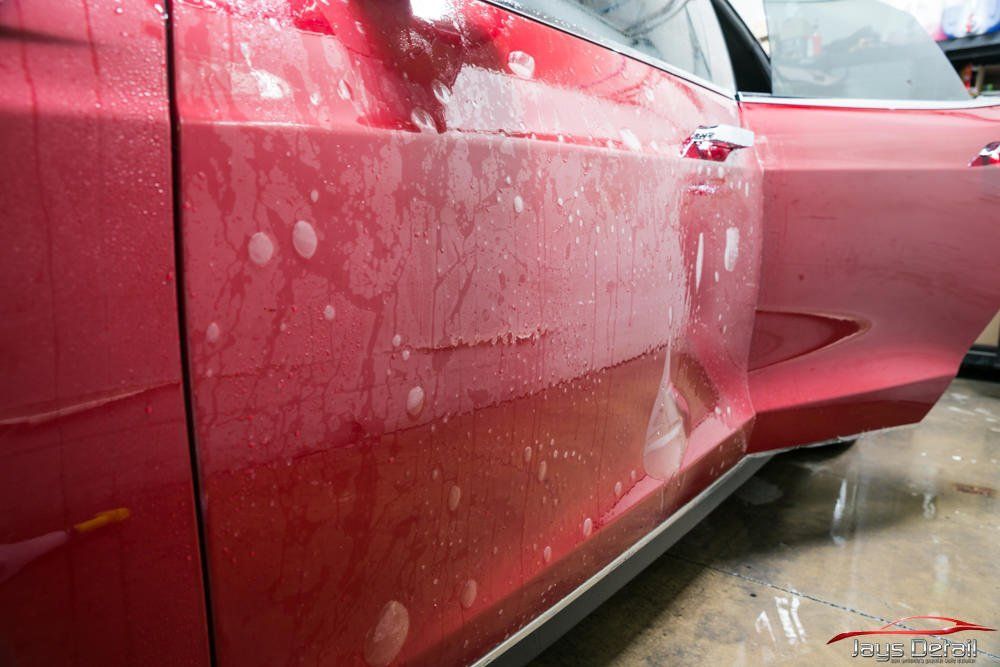 Red car door with clear protective film applied, water droplets visible.