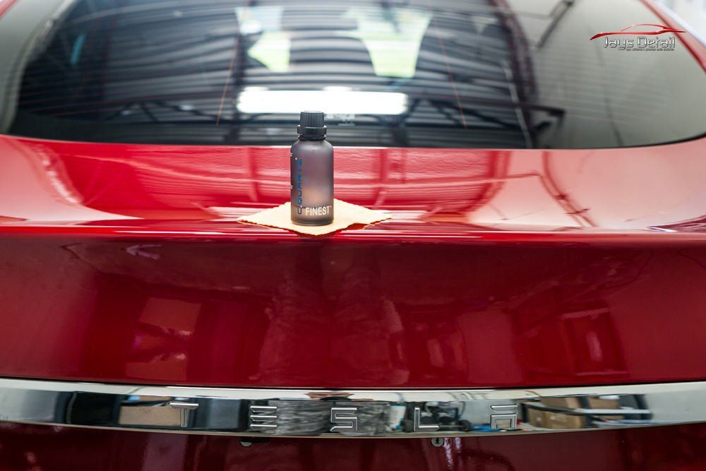 A red car's trunk with a bottle of detailing product, on a microfiber applicator cloth, in an indoor setting.