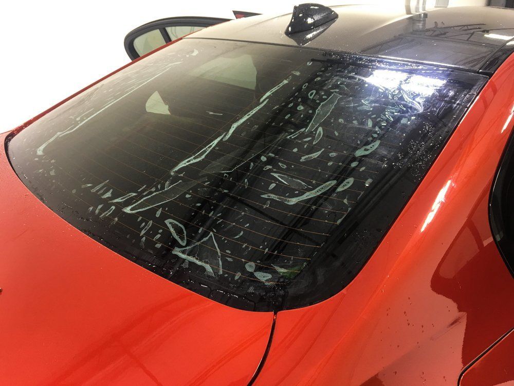 Red car's rear window with clear film being applied; bubbles visible on the surface.