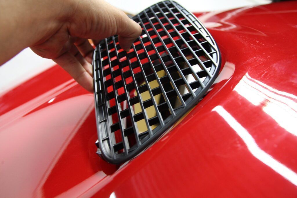 Hand placing a black grill onto a red car's hood vent.
