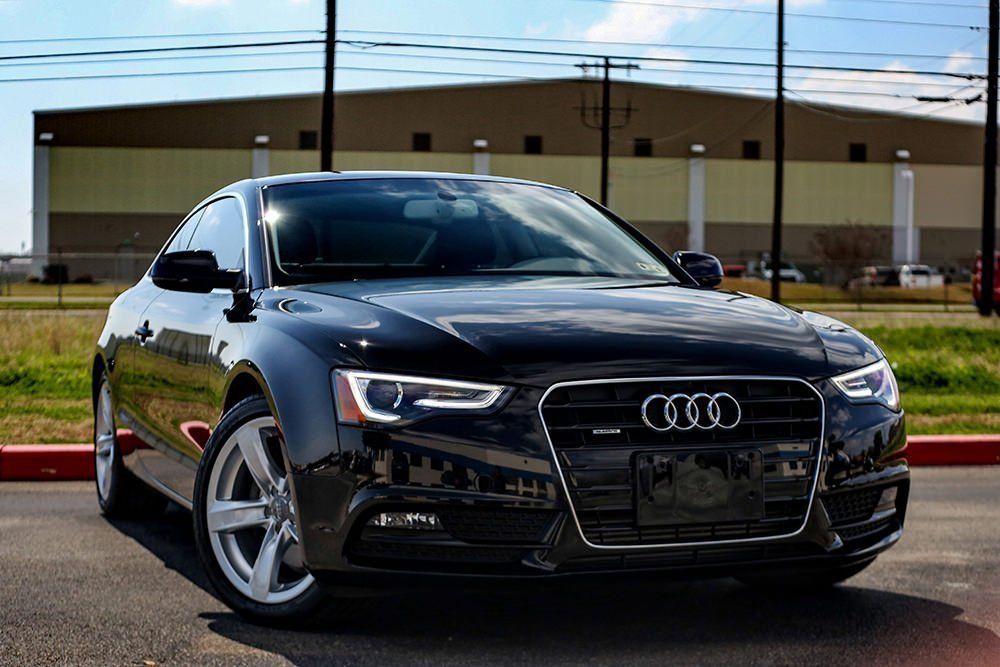 Black Audi A5 coupe parked on a paved surface with a large building in the background.