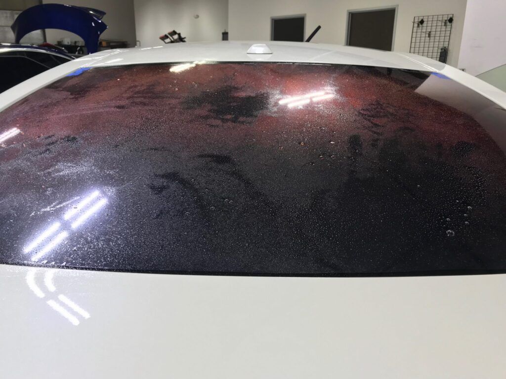 White car's rear window with dark tint, water spots, and reflection of light.