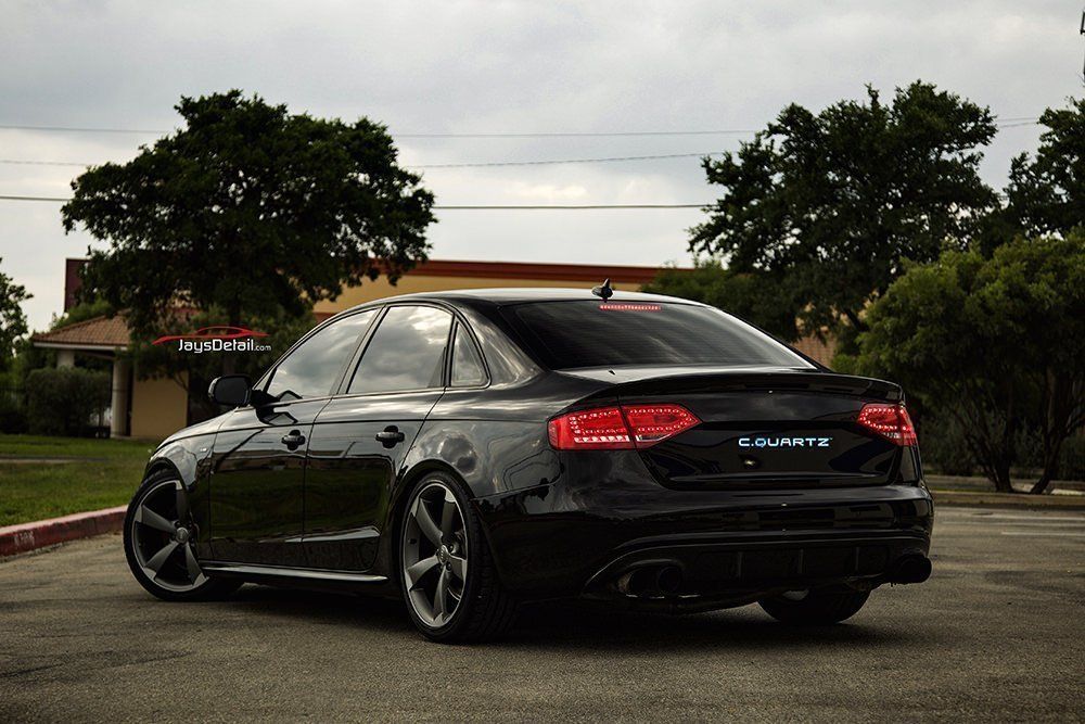 Black Audi sedan parked on asphalt, tinted windows, dark rims, with trees and buildings in background.