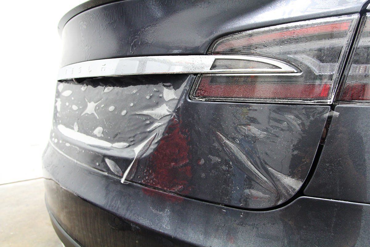 A damaged Tesla rear bumper with peeling film wrap and chrome trim.