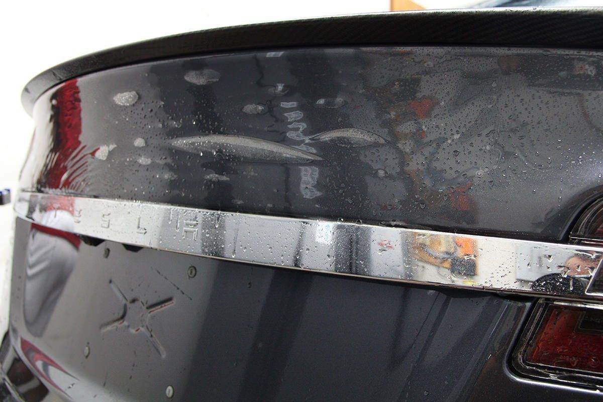 Rear section of a dark car with chrome trim and a black spoiler, wet with water droplets.