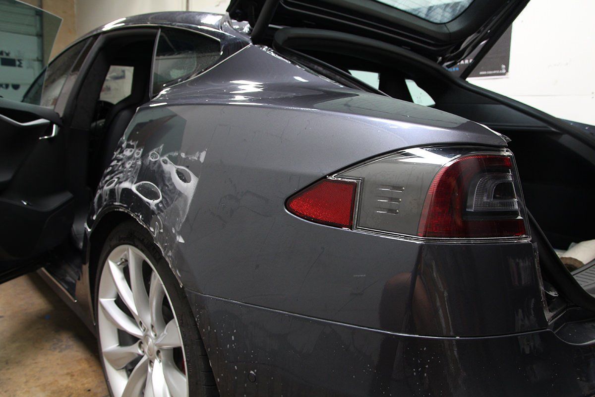 Damaged gray Tesla, rear quarter panel, trunk open, door ajar, garage.
