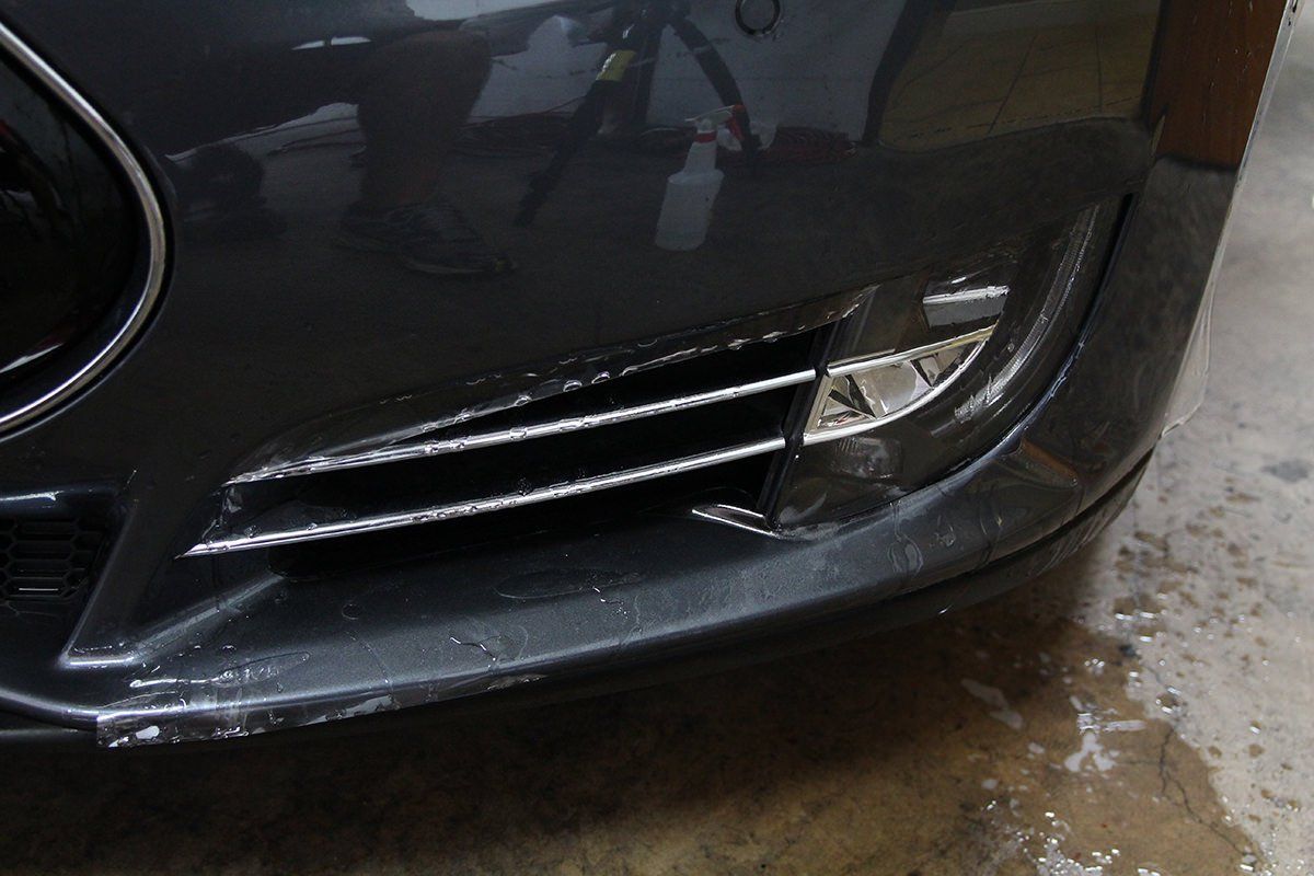 Close-up of a dark gray car's front bumper, showing damaged trim around the fog light.
