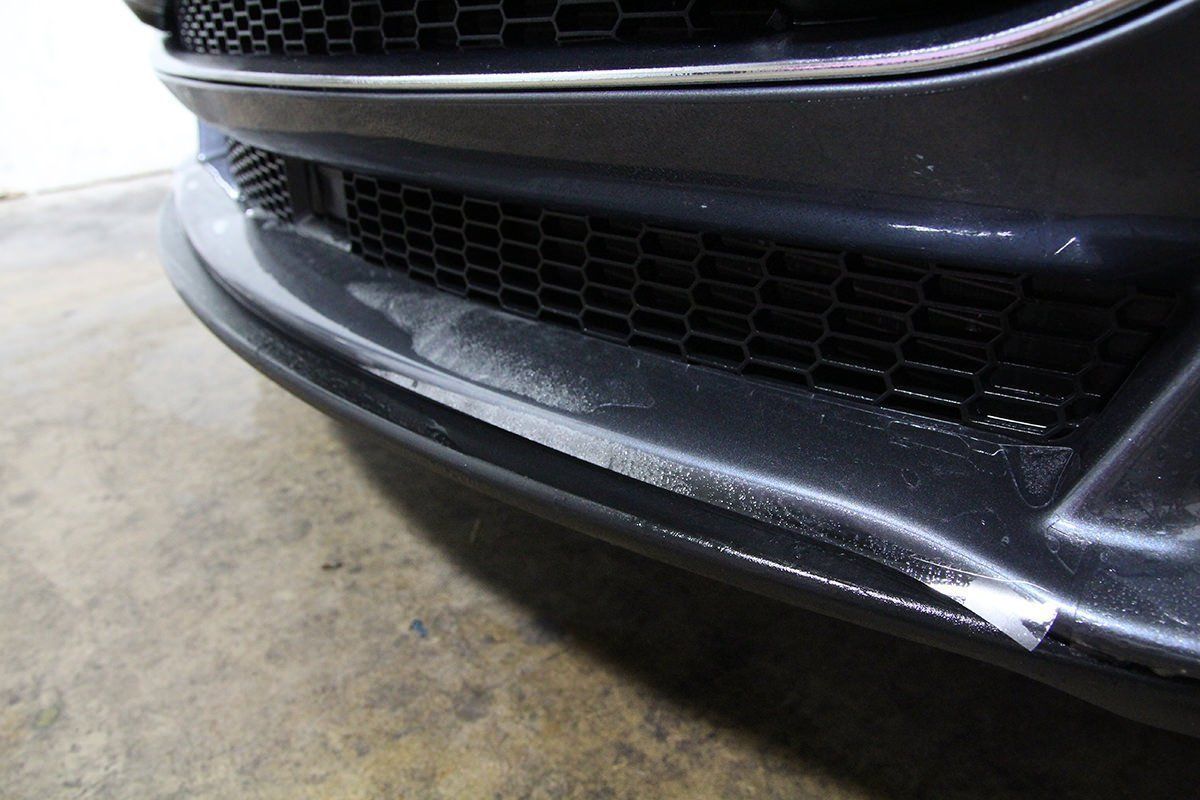 Close-up of a car's front bumper with peeling paint. The bumper is dark grey, showing damage and wear.
