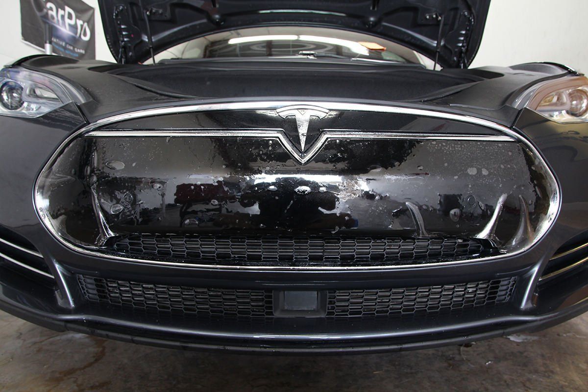 Close-up of a dark gray car's front bumper with damaged trim, showing a vent.