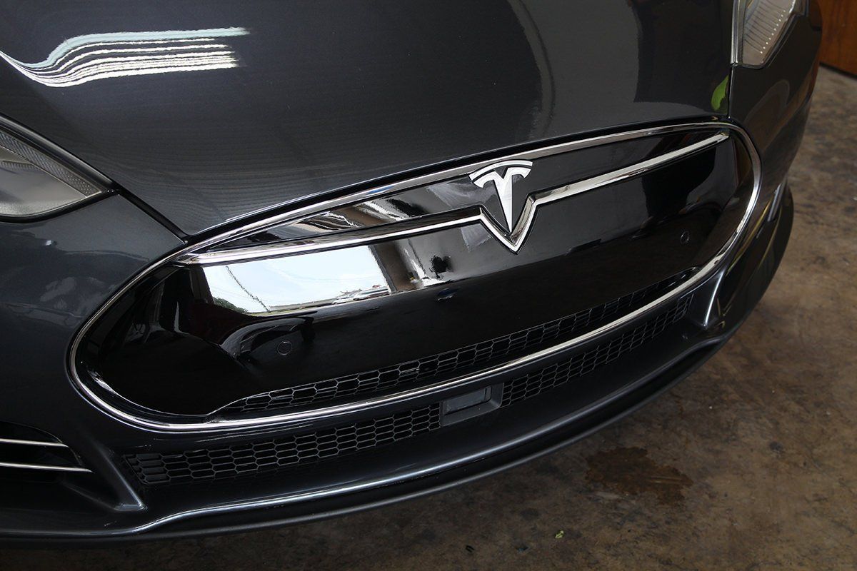 Close-up of a gray Tesla electric car's front. Chrome emblem and grill reflect surroundings.