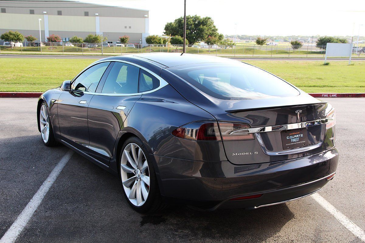 Dark gray Tesla sedan parked in a lot, with a tree and buildings in the background.
