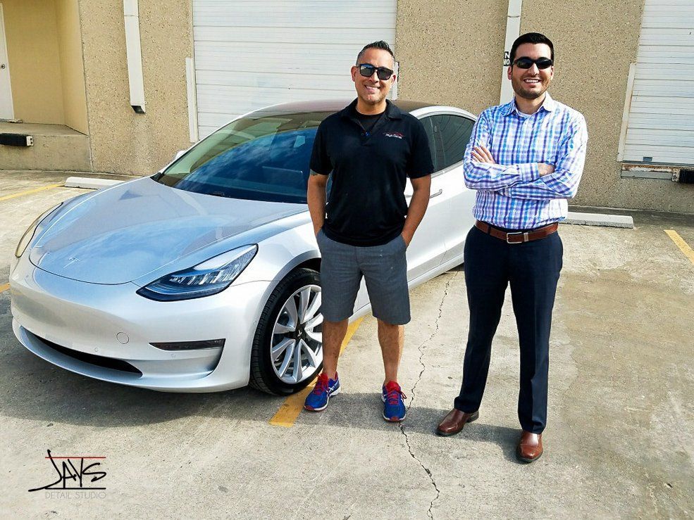 Two men pose next to a silver Tesla. One man wears a black shirt and shorts, the other, a blue button down and slacks.