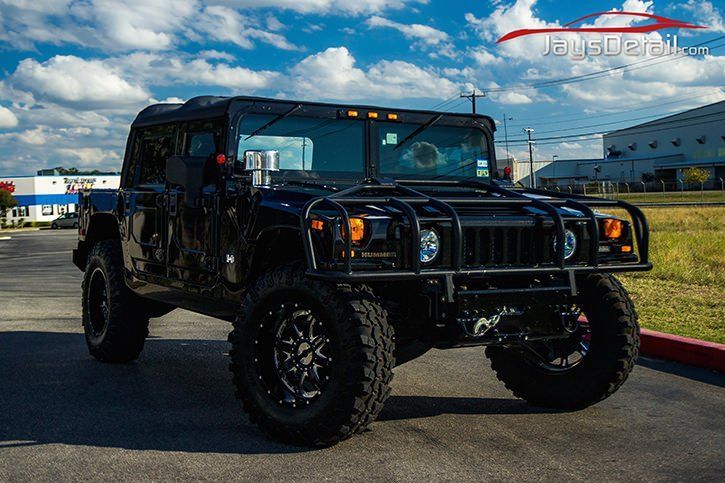 Black Hummer H1 with large wheels parked on pavement, in front of a building on a sunny day.