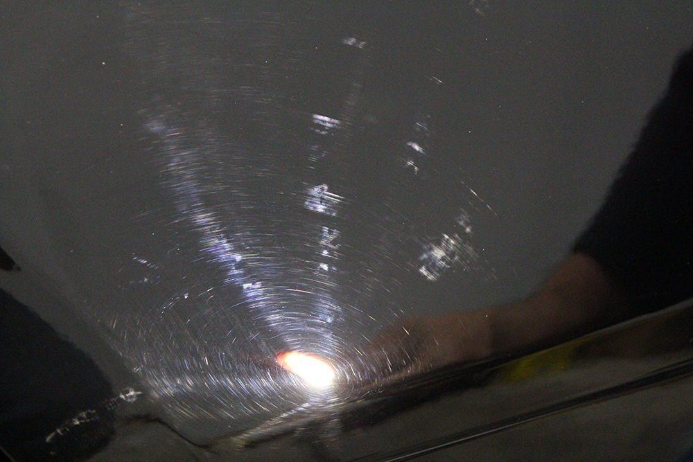 Person polishing a black car, showing swirl marks under a focused light.