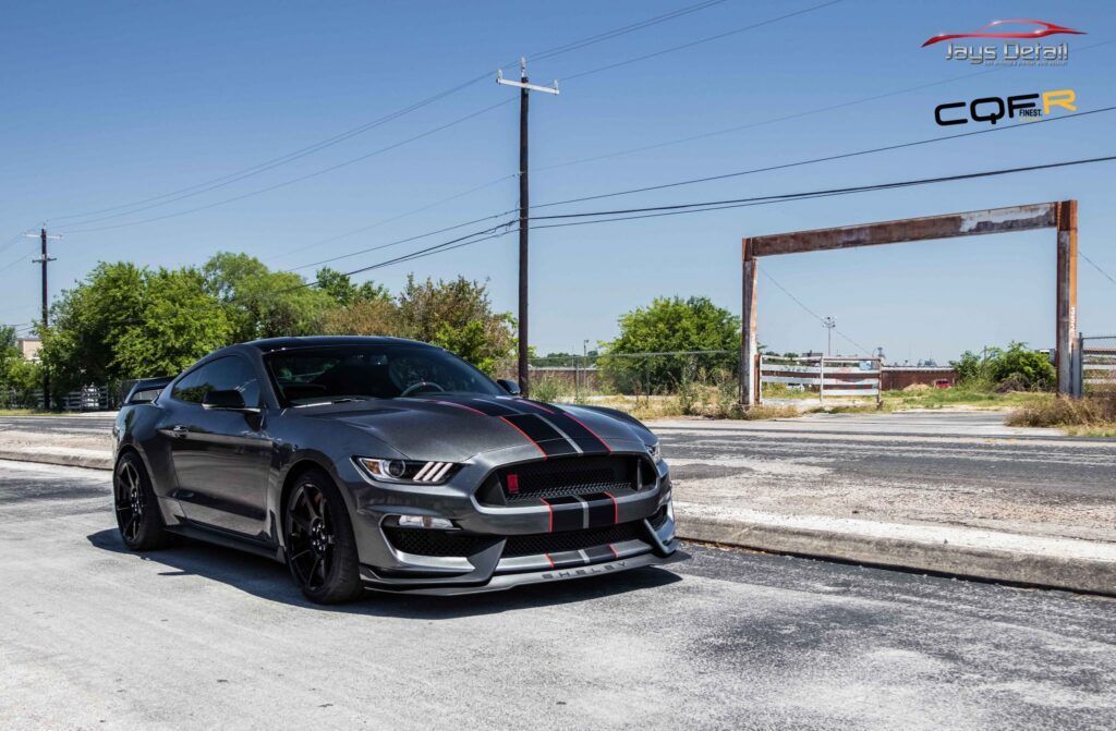 Gray Ford Mustang with black racing stripes parked on a road with a sunny outdoor background.