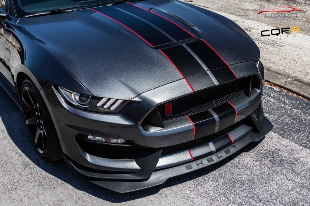 A gray Shelby Mustang sports car with black racing stripes and red accents parked on a paved surface.