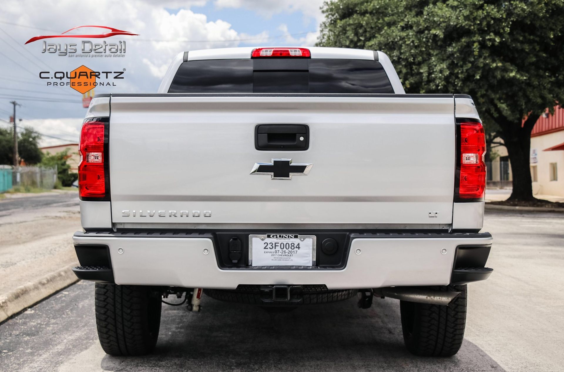 Silver Chevrolet pickup truck, rear view, parked on street. Black bumper, taillights, license plate, and Chevrolet logo visible.