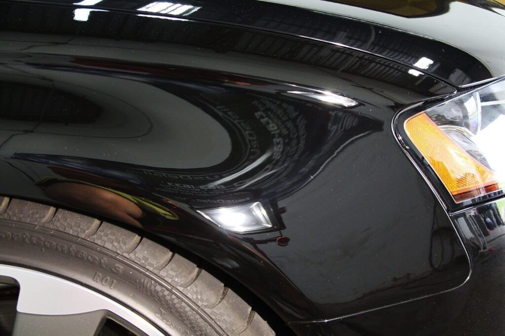Black car fender reflecting light and a portion of a wheel and headlight.